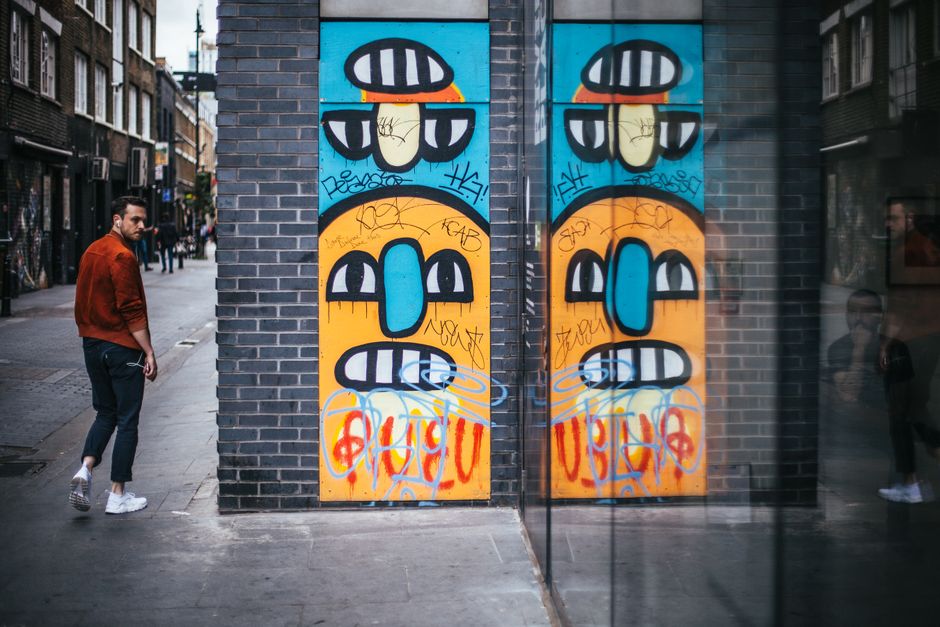 A man with dark hair and a beard, wearing a burnt orange jacket, dark jeans, and white sneakers, stands on a city sidewalk next to a brick wall. The wall features a large, colorful graffiti mural with two symmetrical abstract faces, each with blue noses, orange mouths, and white teeth, topped with black and white striped hats against a bright blue background. The graffiti also includes black tags and orange and red spray-painted words at the lower section of the mural. In the background, the urban street scene shows a narrow pedestrian pathway lined with multi-story buildings, some with brick facades and modern windows. The street appears lively, with a few pedestrians visible further down the sidewalk, and the overall lighting suggests daytime with natural light illuminating the scene. This setting aligns with urban areas where external wall art is common, and the scene reflects a typical city environment often associated with independent creative expressions, paralleling the context of private waste handling and street-level visibility often seen in rubbish removal or clearance services.