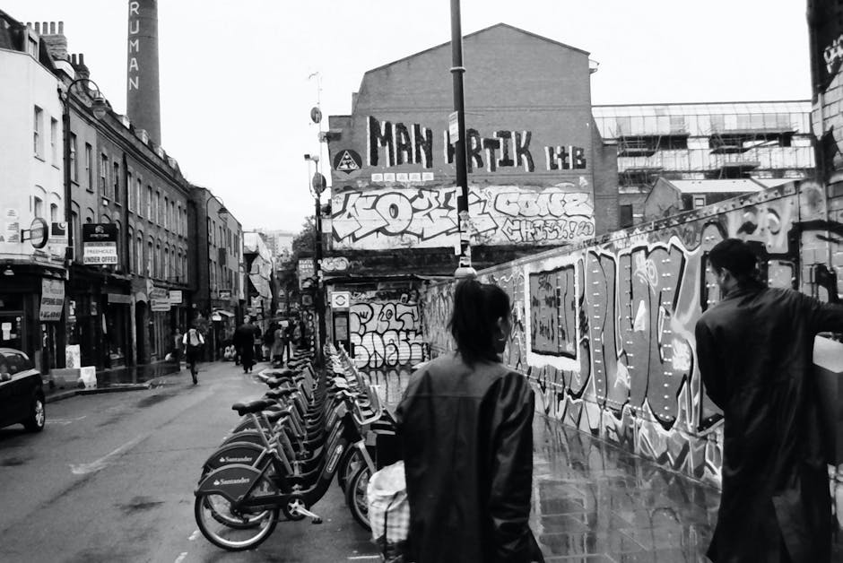 A black and white street scene showing a row of parked bicycles along a paved sidewalk in the foreground. Behind the bikes, two pedestrians, a woman and a man, are walking and standing near a graffiti-covered wall with large, stylized tags and throw-ups painted in various shades of gray, black, and white. In the background, multi-story buildings with shopfronts and signage line the street, with some storefronts displaying awnings and window displays. The street appears slightly wet, reflecting some of the pedal bikes and pedestrians. A lamp post stands near the graffiti wall, and in the upper part of the scene, a large building with a sign that reads 'MAIN ARTIK LAB' is visible. The overall scene depicts an urban environment with street art, casual pedestrian activity, and a roadside setting aligned with alternative waste management or rubbish removal services such as those provided by Rubbish Clearance Shoreditch, with elements of the environment possibly requiring cleaning or rubbish collection.
