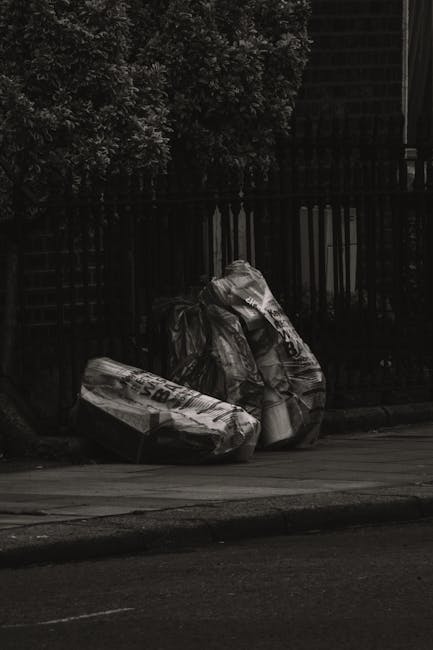 Two large black plastic garbage bags filled with waste material are positioned on the curbside pavement in front of a dark metal fence, with dense foliage and trees visible behind the fence. The bags appear to be tightly knotted at the top, with some creases and surface textures indicating thick plastic, and areas showing slight reflective sheen. One bag is slightly leaning against the other, both placed close together on an asphalt roadway with a curb edge. The scene suggests the bags are awaiting collection, representing private waste removal services such as those provided by Rubbish Clearance Shoreditch, in an urban environment where independent rubbish disposal is common. The lighting appears natural and somewhat subdued, consistent with ambient daytime or early evening conditions. The environment is clean, with no visible rubbish scattered around, and the background fence and greenery create a quiet, residential street context.