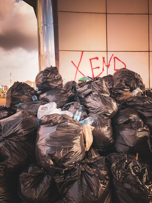 A large pile of black plastic rubbish bags, some torn open revealing plastic bottles and other waste, stacked against a modern building with beige tiled exterior panels. The bags are piled in an outdoor urban setting, partially illuminated by natural light with a cloudy sky in the background. There is a red spray-painted graffiti tag on the wall behind the rubbish, reading 'XEND'. The scene illustrates an example of unmanaged waste accumulation that could be addressed through private rubbish clearance services such as those offered by Rubbish Clearance Shoreditch, providing an alternative to council-led rubbish disposal methods within a city environment. The bags are in various states of fullness, with some tied tightly and others loosely gathered, and the bottles in the scene suggest mixed waste materials. Overall, the composition emphasizes the unmanaged waste clutter typical of outdoor city refuse, suited for professional rubbish removal specialists to handle efficiently.