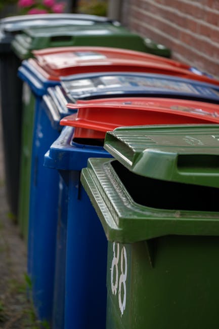 A row of large wheelie bins lined up outdoors along a pavement, with the front green bin closest to the camera and the others in blue, red, and dark green extending into the background. The bins are made of plastic with textured lids, some of which are partially open, revealing the interior space. They are positioned against a brick wall on the right side, suggesting a residential or commercial waste collection area. The environment appears to be an alleyway or driveway, with natural daylight illuminating the scene. The image relates to rubbish removal services provided by Rubbish Clearance Shoreditch, illustrating an example of private waste collection or on-site rubbish storage prior to collection, highlighting the need for efficient waste management solutions in urban settings.