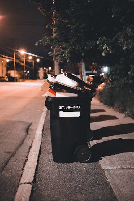 A row of large wheelie bins lined up outdoors along a pavement, with the front green bin closest to the camera and the others in blue, red, and dark green extending into the background. The bins are made of plastic with textured lids, some of which are partially open, revealing the interior space. They are positioned against a brick wall on the right side, suggesting a residential or commercial waste collection area. The environment appears to be an alleyway or driveway, with natural daylight illuminating the scene. The image relates to rubbish removal services provided by Rubbish Clearance Shoreditch, illustrating an example of private waste collection or on-site rubbish storage prior to collection, highlighting the need for efficient waste management solutions in urban settings.